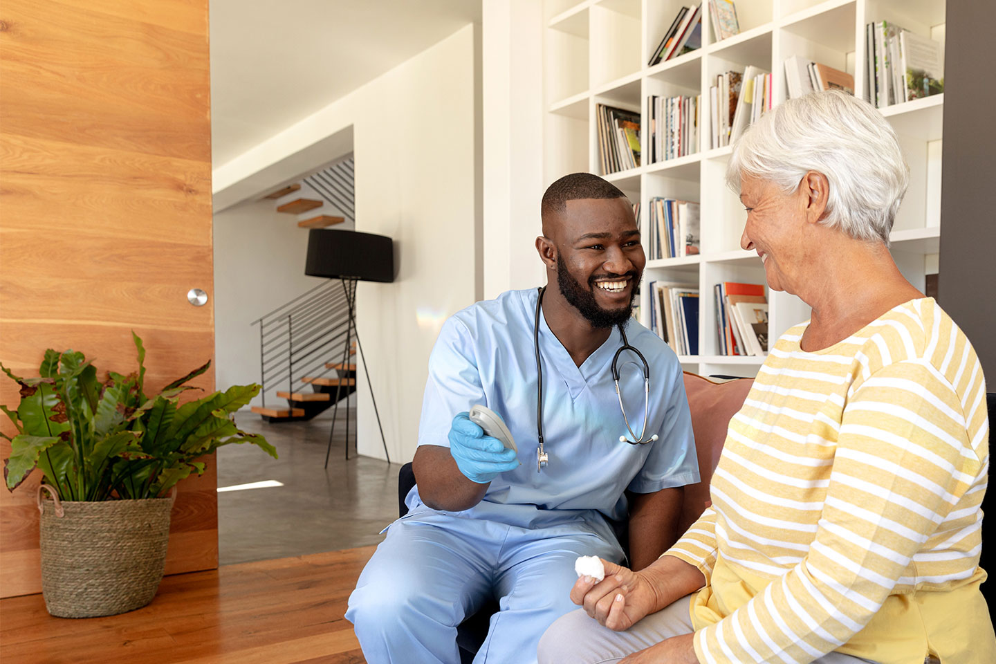 Nurse in blue scrubs showing an older woman wearing a white and yellow striped shirt the results from her blood prick test on a small gray electronic device.